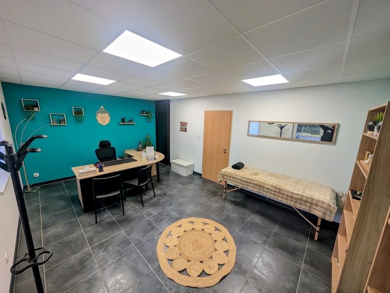 Le cabinet de cartomancie de Carmela à Marckolsheim : bureau en bois devant un mur turquoise décoré d’un bouddha, table de soin et tapis rond en jute